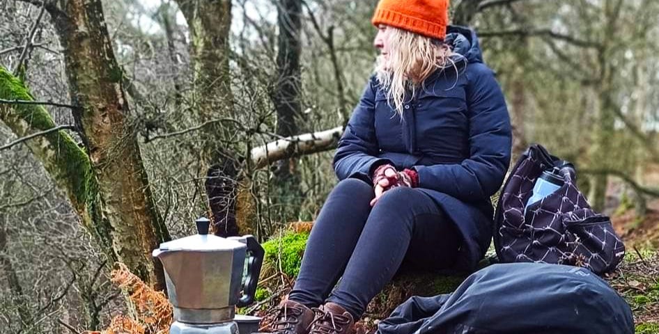 A woman with blonde wavy hair wearing an orange bobble hat, dark blue coat, bvlack leggings and brown walking boots is sitting on a log in woodland. There is a coffee pot on a small stove in front of her with two cups and a rucksack.