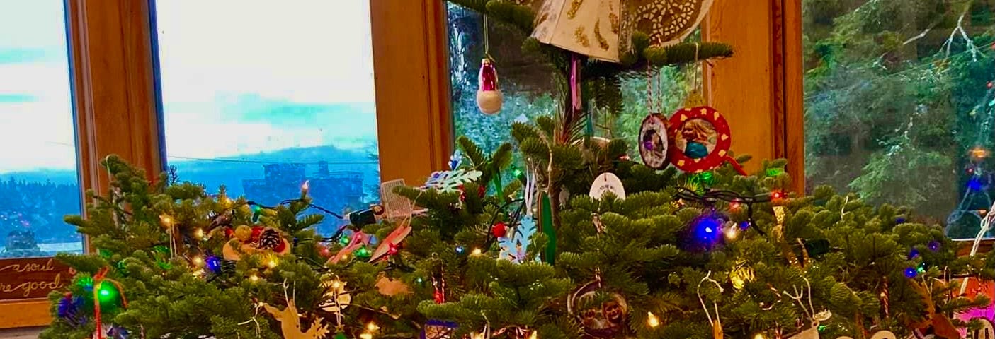 A misshapen Christmas tree is framed by a window. The tree is decorated with hand made decorations, a string of lights and a Christmas angel sits on top.