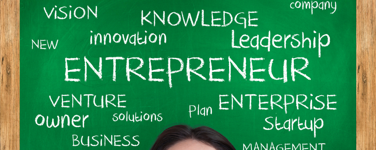 The top half of a young woman’s face looks up at a chalkboard with entrepreneur-related words written all over it in chalk.