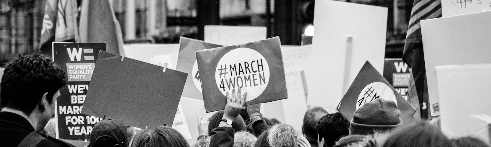 black and white photo of a contemporary political march for women; a crowd of mostly women holding pro-women signs, shot from the back