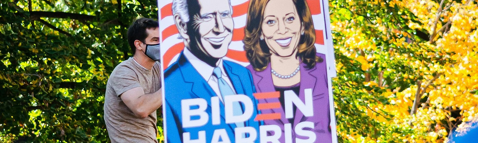 Photo of a man with a COVID mask over his mouth holding a “Vote Biden-Harris” sign, while sitting high atop a stop light.