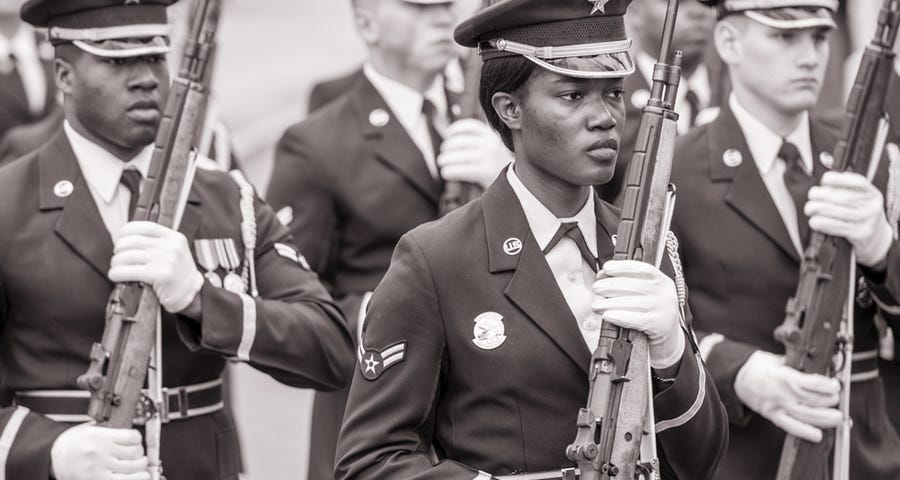 United States soldiers in dress uniforms holding rifles at a military ceremony. Mix of men and women, blacks and whites.