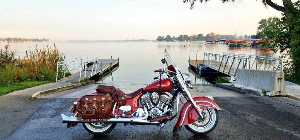 A red Indian Chief motorcycle with dark brown saddlebags, sitting at a boat launch site with a lake in the background.