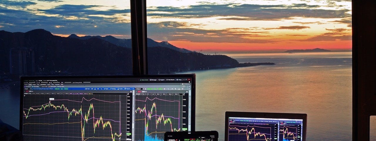 IMAGE: Several computer screens visualizing stock market charts with a wonderful sea sunset view in a window in the background.