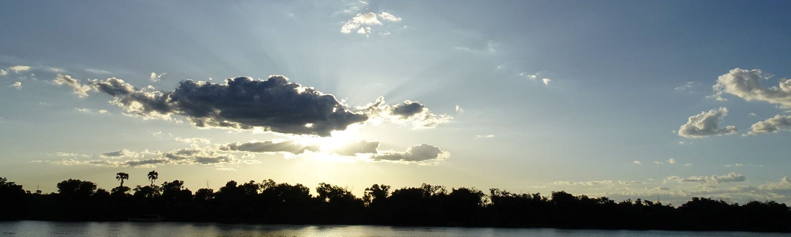 The golden light of the sun setting over a dark river and darker trees, partially behind clouds