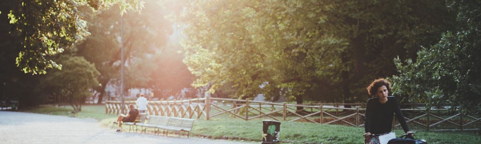 A woman riding a bike in a park with the sun shining through trees behind her.