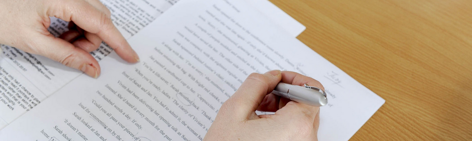 Hands in a sweater hover over a typed piece of paper with edit marks on it all over a light wooden surface. The left hand points at a line while the right hand holds a pen.