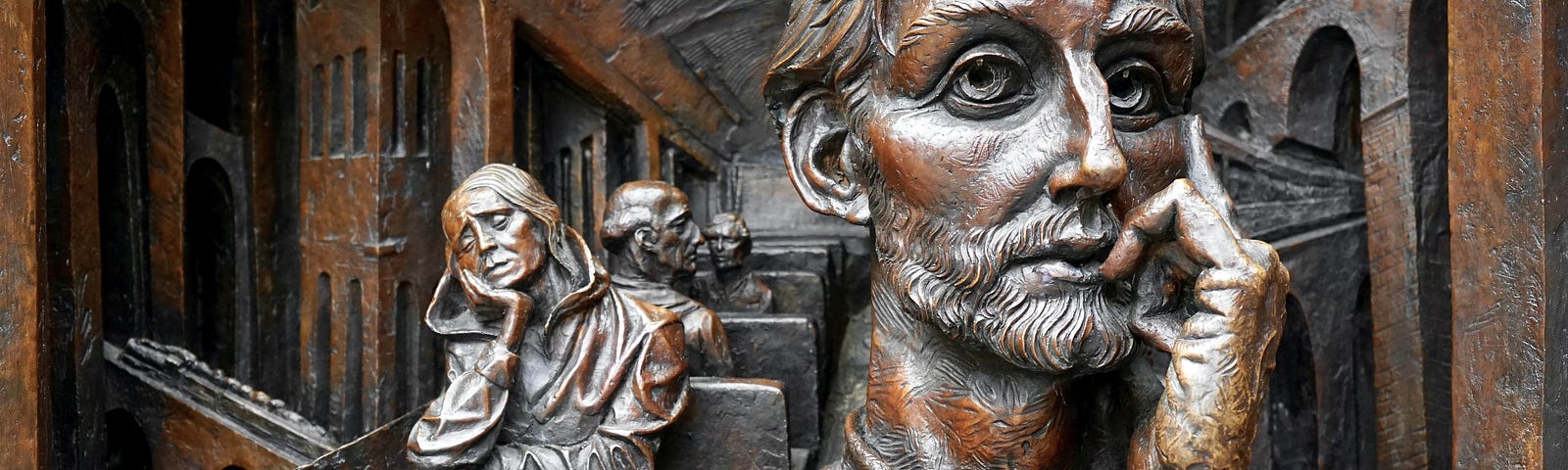 Bronze statue of a man in the foreground sitting in a thinking pose. Behind his right shoulder is a sleeping woman