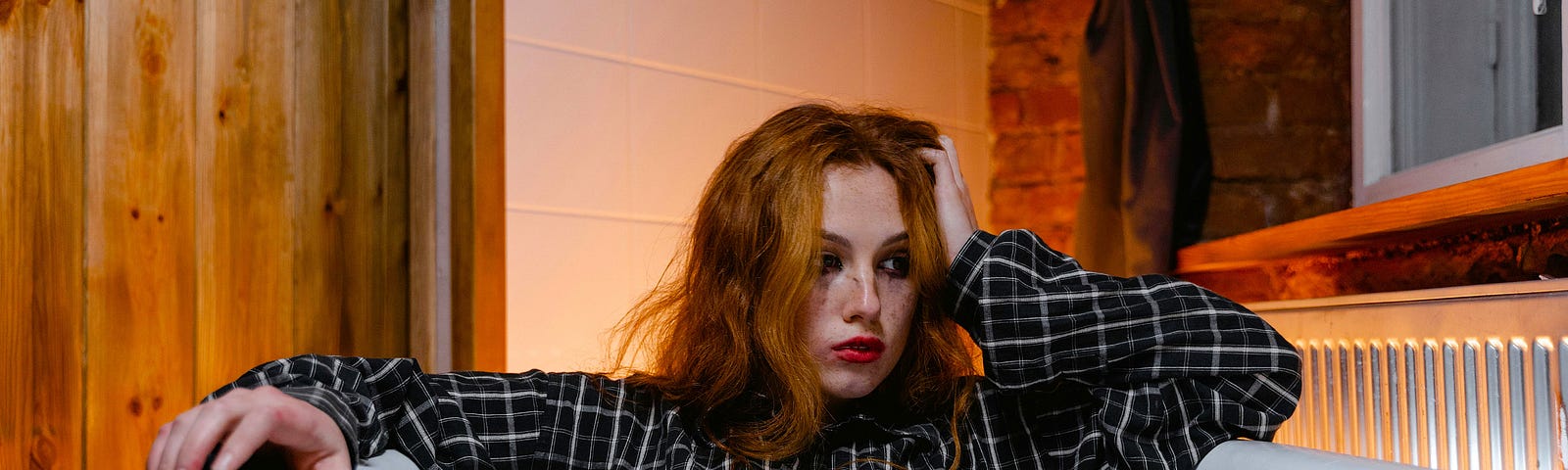 a color photograph of a young person wearing a long dark shirt and pants with fair skin and long red hair looking rather forlorn, sitting fully-clothed in a bathtub full of white milk