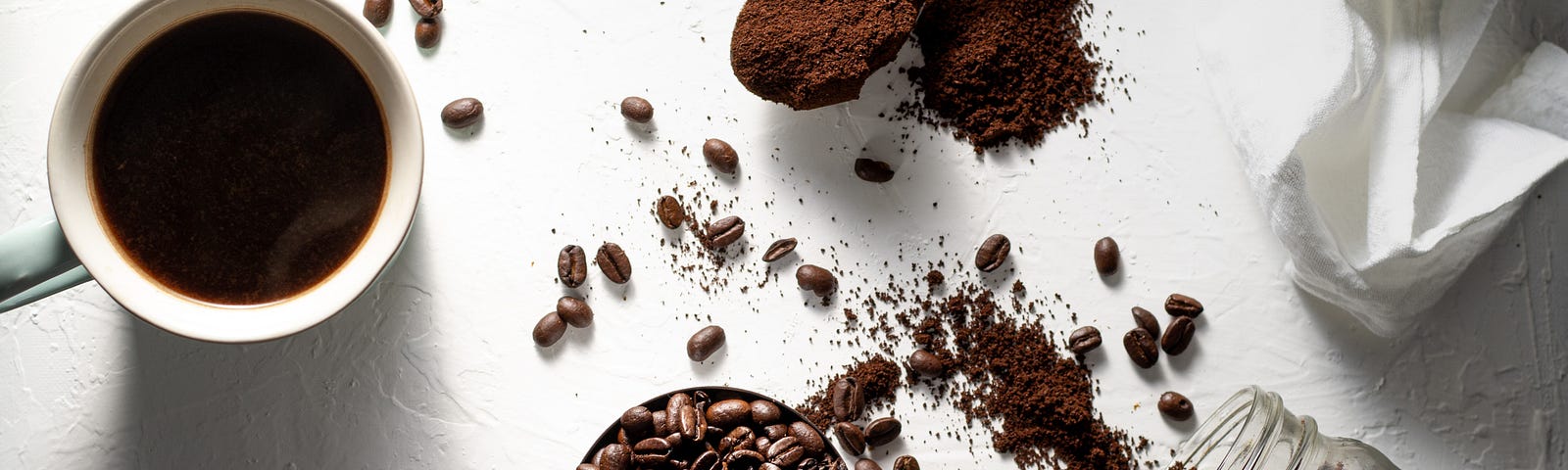 mug of coffee surrounded by whole coffee beans and ground coffee spilled on a white table cloth