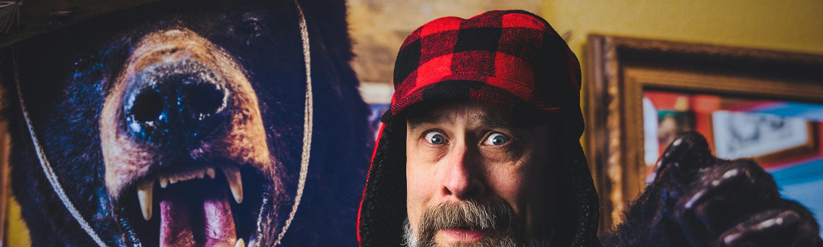 A bearded man with a funny facial expression while standing next to a growling stuffed bear