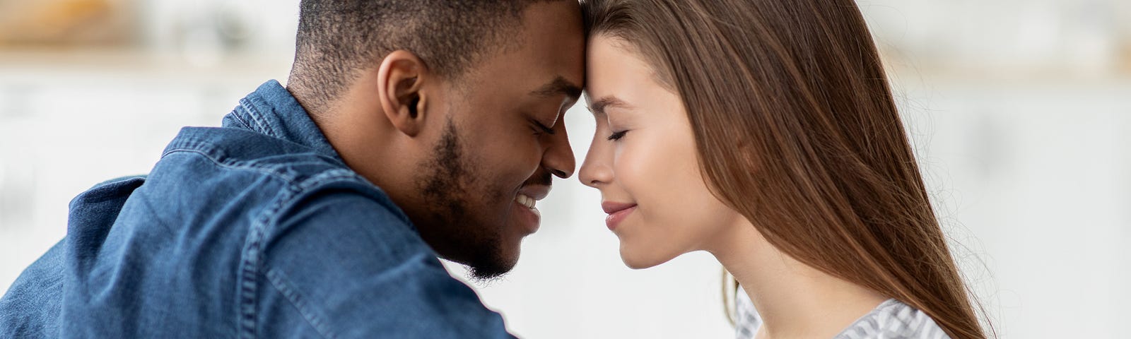 A young black man and a young white woman sit facing each other. Their eyes are closed and their foreheads are touching. Each of them are smiling. It is an intimate moment.