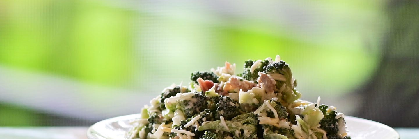 A serving of broccoli bacon salad on a white plate, with an obscured green hued background.