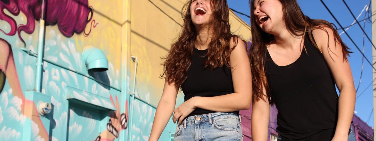 Two brunette women wearing black tank tops and light blue jeans walk down a street and laugh together.
