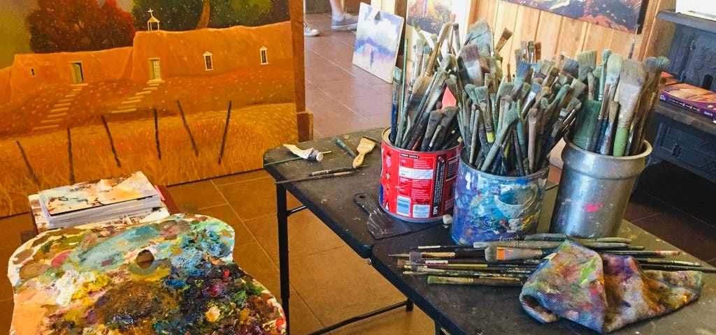 A busy artist’s studio with a dirty color palette and many paintbrushes in cans nearby
