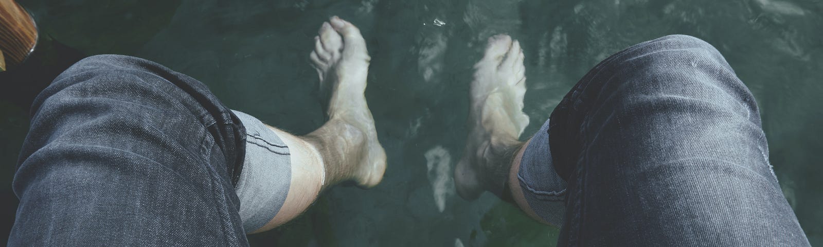 Legs in cuffed jeans sitting on the edge of a wood dock with bare feet in the water below.