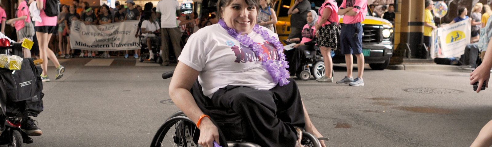 Karen wears a casual outfit and a purple lei, holding a purple noisemaker. She has short brown hair and uses a wheelchair.