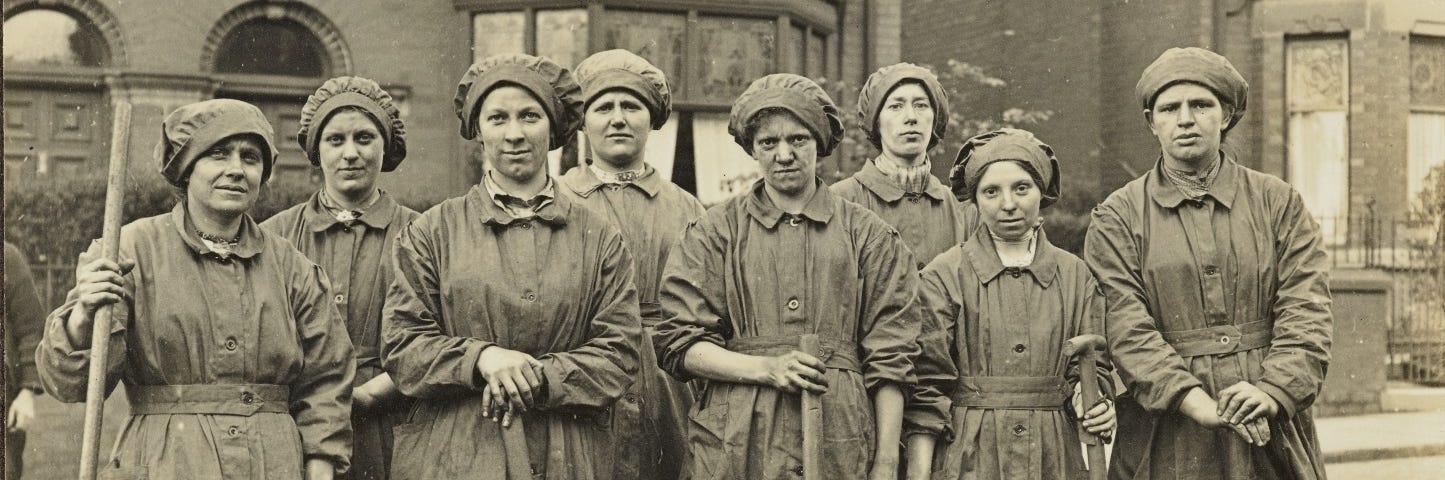 Sepia photograph of 8 female tramway repair workers wearing caps and holding large metal tools, standing in the middle of a cobbled street.