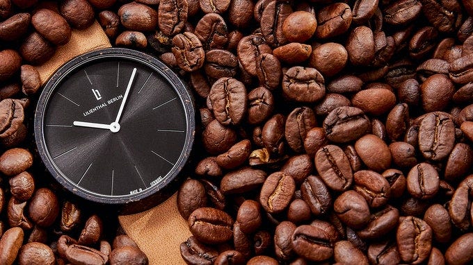 A watch with a light-brown vegan leather strap laying in a pile of coffee beans. The time on the watch shows approximately 10:07, and the watch case is made from recycled coffee grounds.