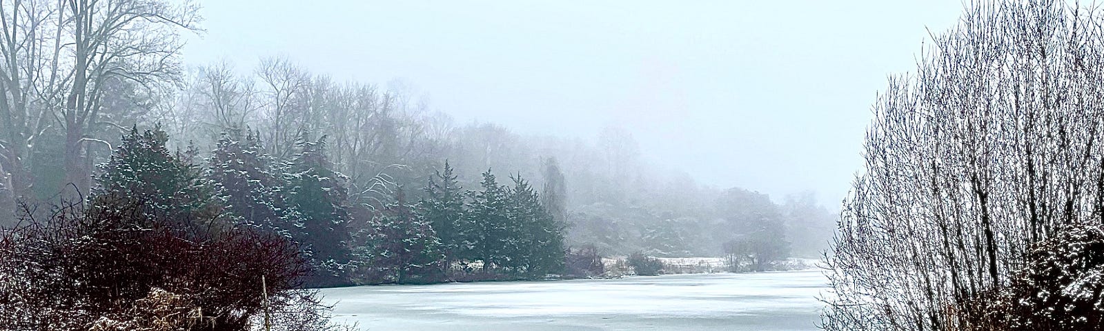 A frigid cold winter scene, a beautiful frozen pond with snow covered evergreen trees on the water’s edge.