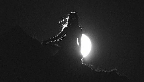 Dark photo of woman standing in front of the moon. Her hair blows to the side and eyes are glowing.