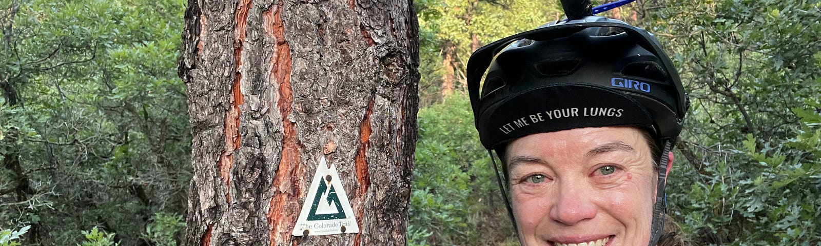 The author, Patty George, smiling in her Team PHenomenal Hope uniform and bicycle helmet, standing next to a tree with the Colorado trail logo. She is wearing a cycling cap with the words “Let Me Be Your Lungs”