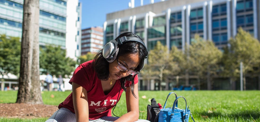 Female student with headphones sitting on grass, writing in a notebook in front of a laptop.