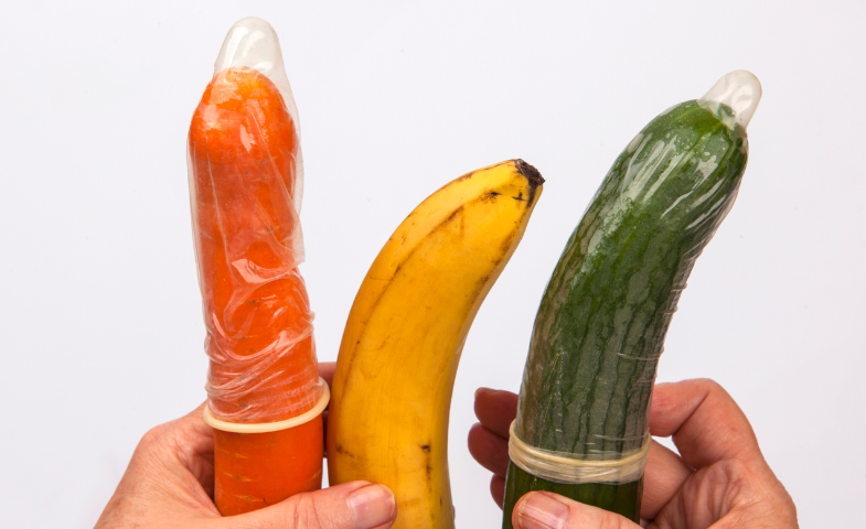 Sobre un fondo blanco, unas manos sostienen una zanahoria, un plátano y un pepino, todos ellos con condones. Crédito: Canva