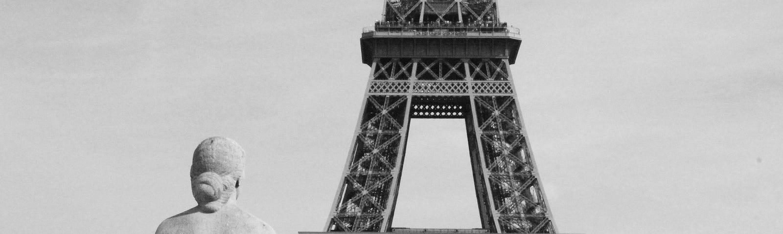 A statue of a woman, looking at the Eiffel tower. She is seated, her is back to the camera. Black and white photograph.