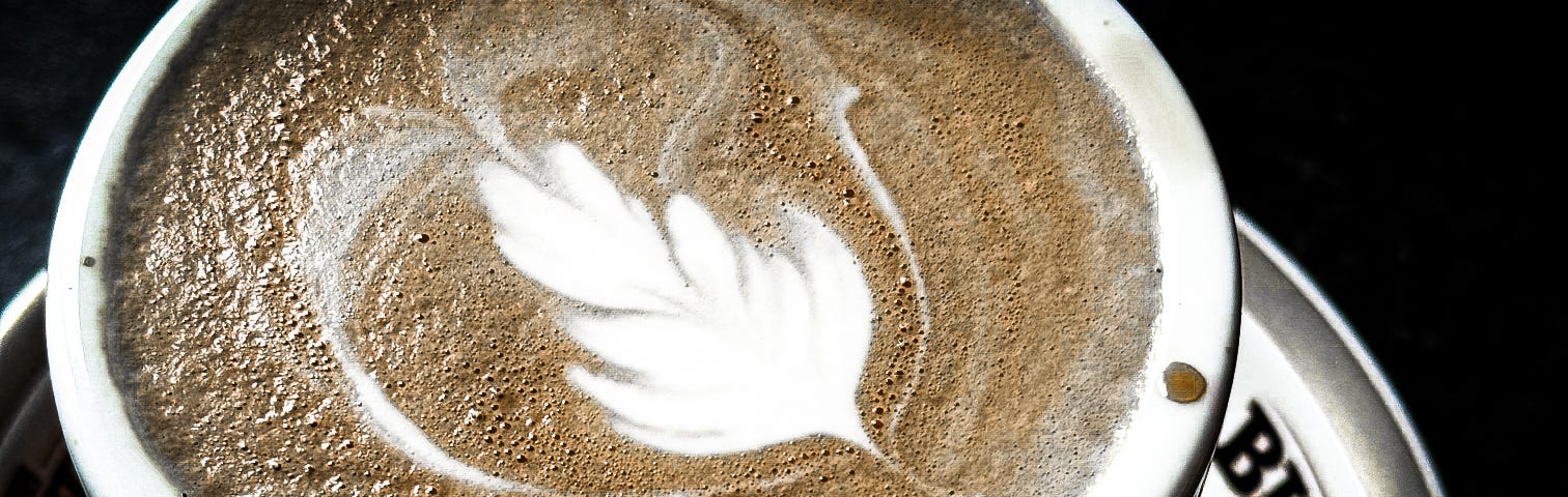 Close up of a flat white coffee with latte art in the shape of a feather, on a “Black and White” branded cup and plate.