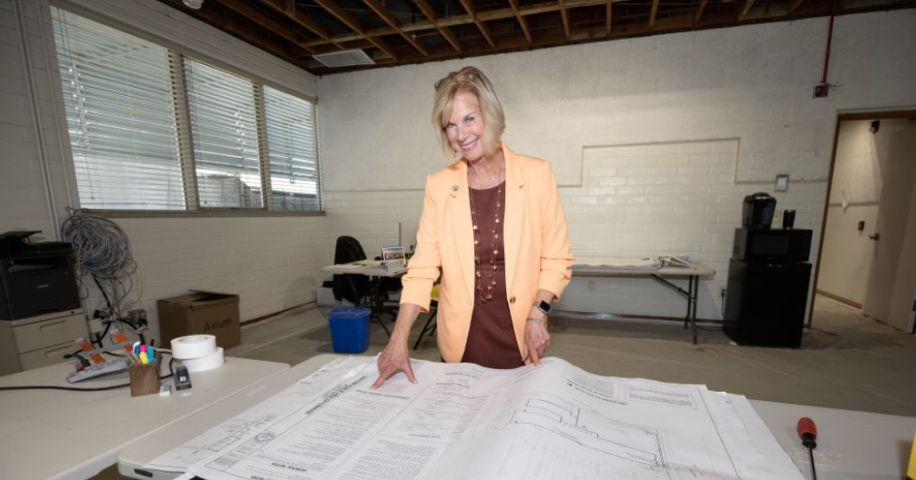 In a room full of tables, boxes, and fans, a woman is in the center of the photo and she is pointing at a long white table. The table has several large papers and appears to be instructions for a possible layout. The slightly older woman is smiling and is wearing a yellow jacket with a brown shirt.