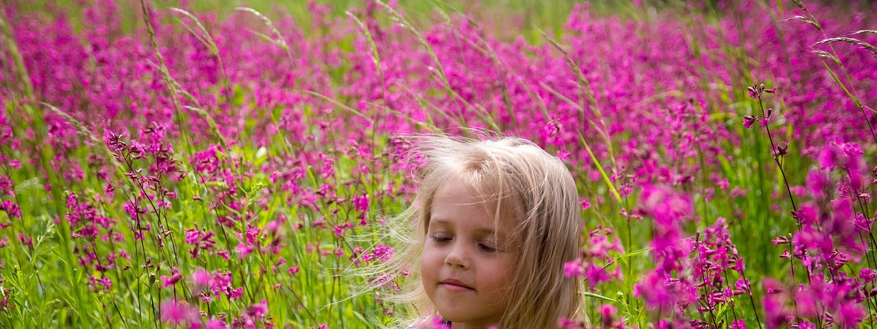 A little blonde haired girl stands in a field of fuschia colored flowers during May. Her eyes are closed and she is dreaming into reality. Every day is a heyday for her with her child’s eye view of the world and innocent wisdom.