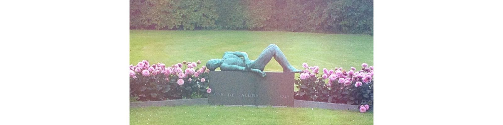A verdigris bronze figure lying on a low, polished stone plinth, pink dahlias at either side, green cemetery lawn behind, looks like a misty, late afternoon