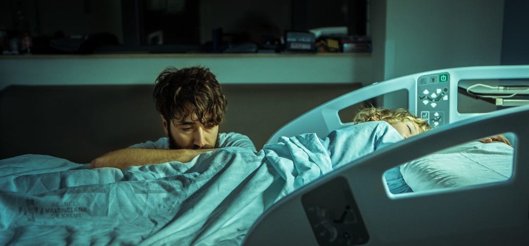 A parent kneels beside a sick child’s hospital bed with a concerned look.