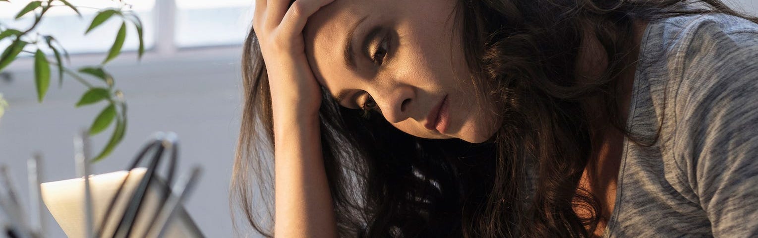 A millennial woman who feels depressed sits at her desk, clutching her forehead and looking at her phone.