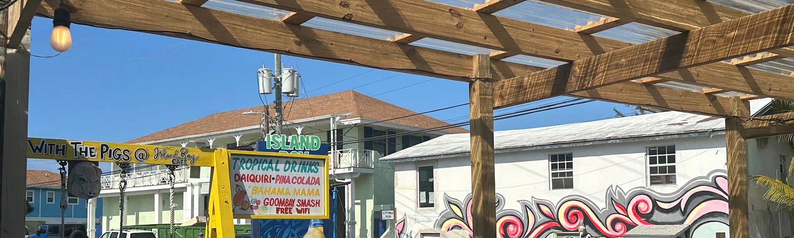 Wood pergola over picnic tables and chairs. A yellow sign — “Island Bar” with a list of a few cocktails. A colonial-style building is at the back of the parking lot with another plain white two-story building next to it. There is pink and grey street art on the white building, that says “Heel 2 Toe”