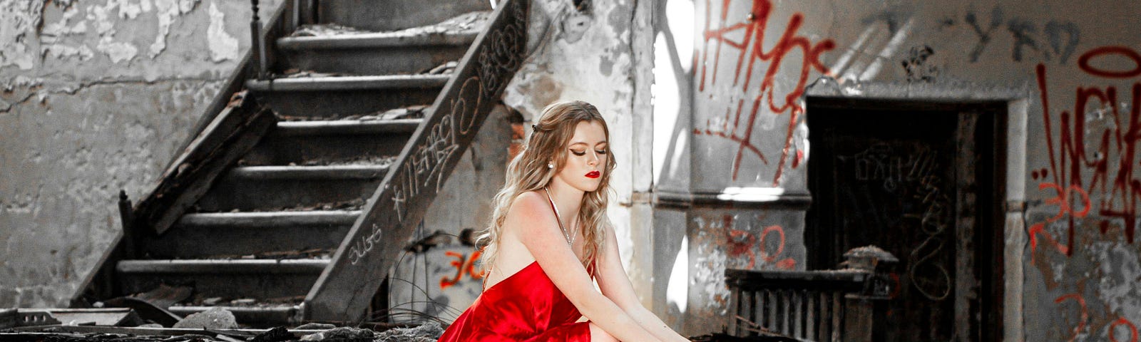 A woman in a red dress is sitting on the stairs of an abandoned building. She’s holding a crown while casting her gaze downward.