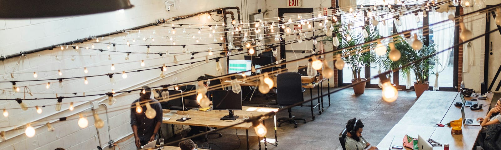 Plunging view of people sitting in front of computer monitors at a coworking space, with light garlands crossing the ceiling.