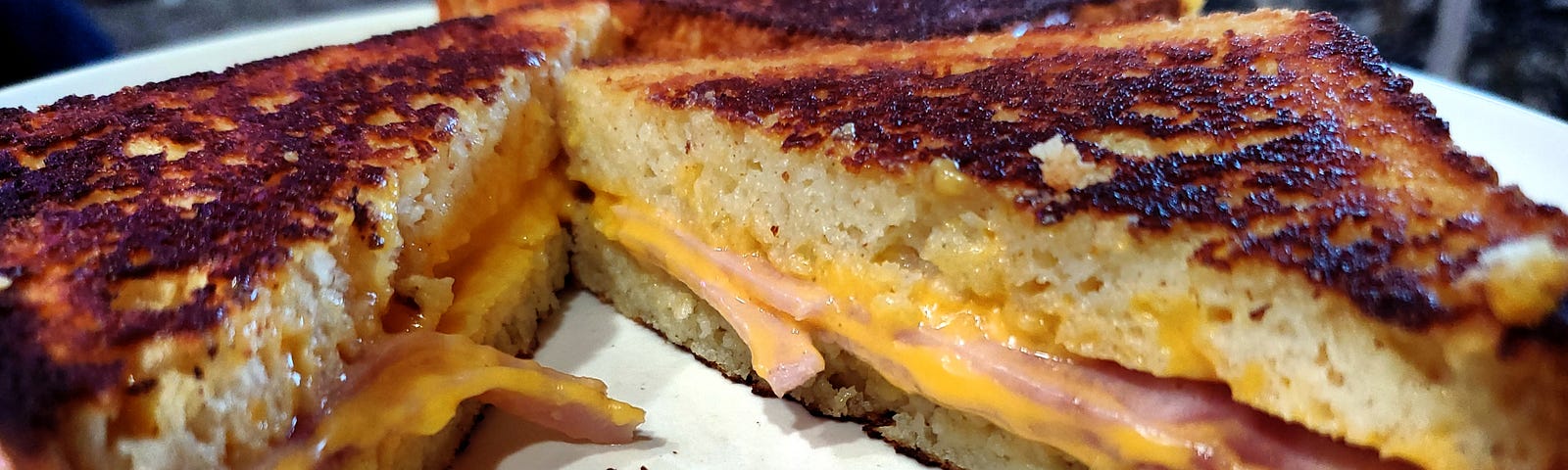 A toasted sandwich with melted cheese and ham is cut diagonally and placed on a white plate. It’s grilled to a golden-brown color and appears crispy on the exterior, with the cheese oozing out from the middle.