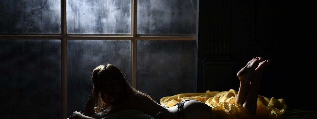 Image shows a nighttime scene in a bedroom, with moonlight on the other side of a window, and a woman wearing only a string lying on her front, looking at the window, her head propped up on her hand and her legs bent, feed pointing up.