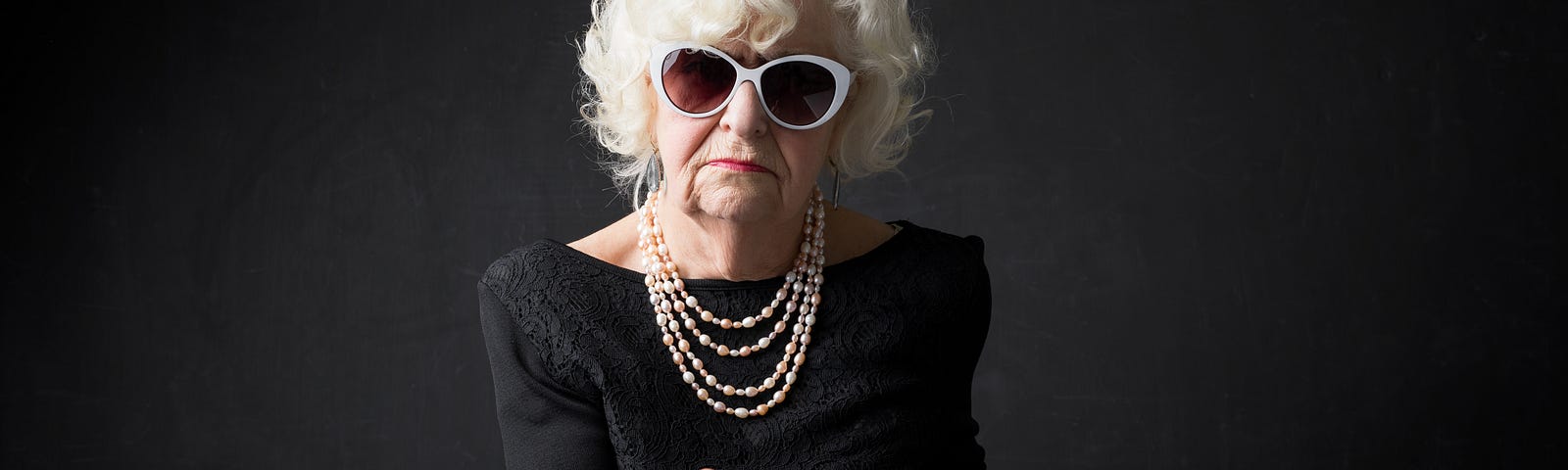 A wealthy elderly woman wearing pearls and sun glasses looks straight into the camera and gives the finger.