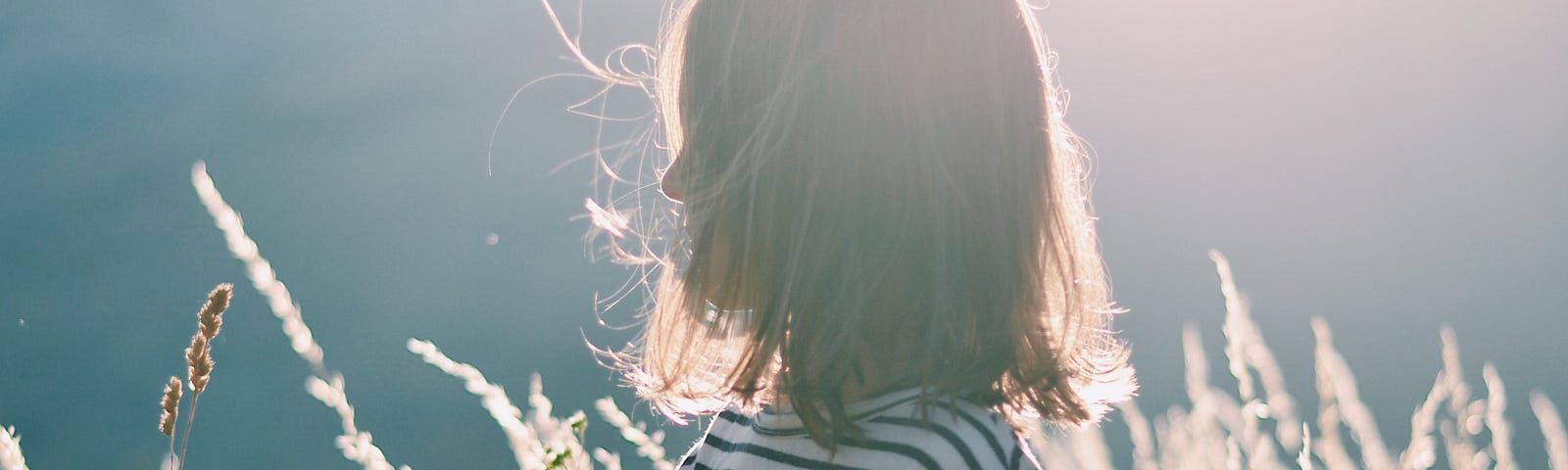 Little girl with dark hair wearing a stripped dress in a field touching the piece of wheat.