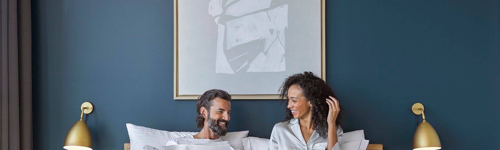 A man reading the newspaper, and a woman working on her laptop in bed