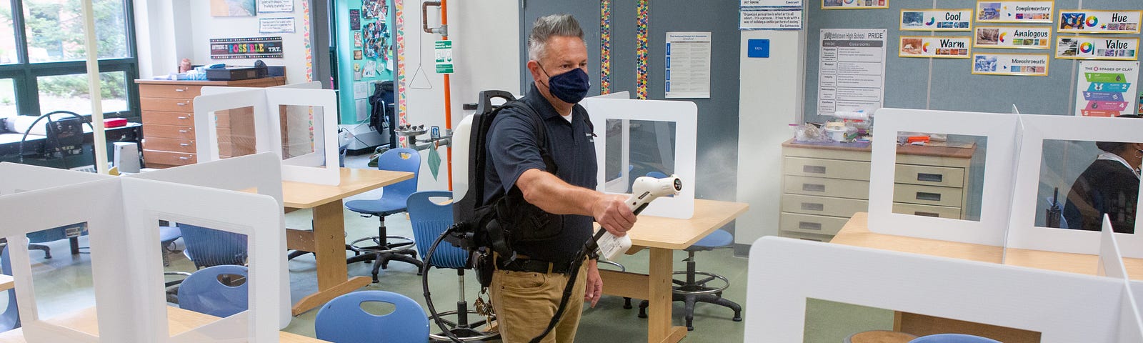 An adult man wearing a face mask sprays disinfectant in an empty classroom.