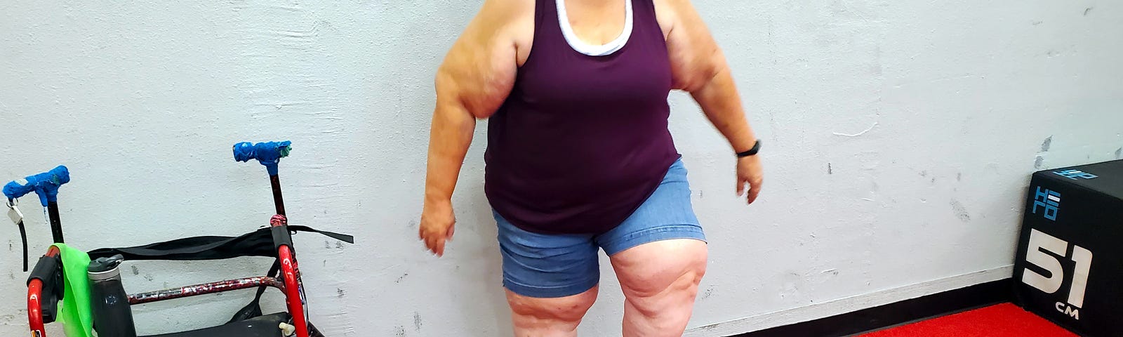 Older fat woman wearing a burgandy tank top and blue shorts stepping up on an exercise step next to a red wheeled walker