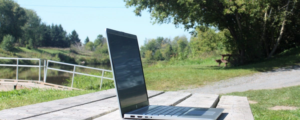 A laptop computer on a picnic table in a park-like setting by the water.