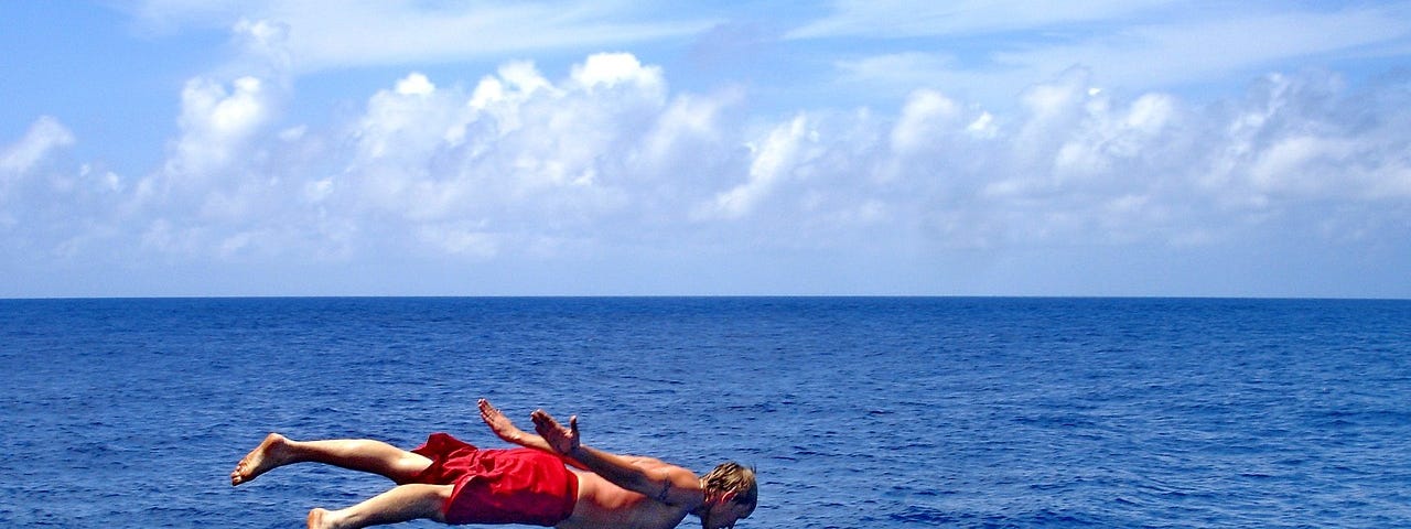 A man pictured suspended over the ocean under a partly cloudy blue sky, the camera caching him in the middle of his dive.