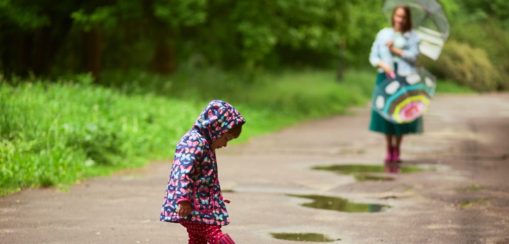 Photo of mom standing behind with umbrellas while her daughter plays in a pothole of water after a rain fall.