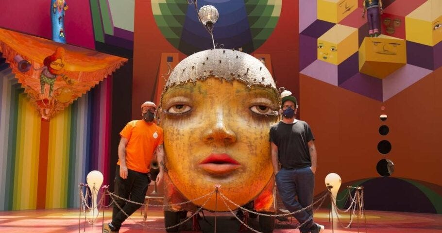 Os Gêmeos na primeira sala da exposição, a qual conta com uma escultura ao centro e pinturas coloridas nas paredes.