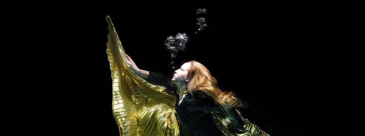 An underwater female model wearing a short black dress and material floating around her like gold wings.